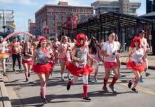 A group of participants in red tutus and festive outfits run down a city street during Cupid’s Undie Run, with buildings and spectators visible in the background.