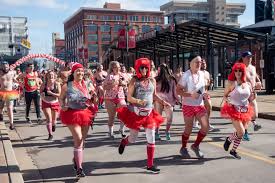 A group of participants in red tutus and festive outfits run down a city street during Cupid’s Undie Run, with buildings and spectators visible in the background.