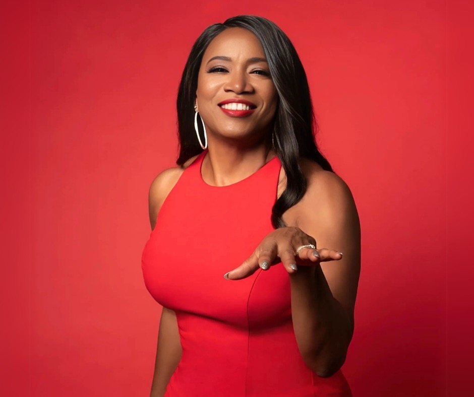 Portrait of singer Eugenie Jones against a bold red background, wearing a sleeveless red dress and smiling confidently toward the camera. She gestures with one hand extended, conveying warmth and charisma, with studio lighting highlighting her expressive stage presence.