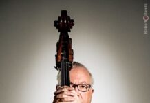 Portrait of a man holding an upright bass vertically in front of his face, standing against a neutral background, with only his eyes and arms visible around the instrument.