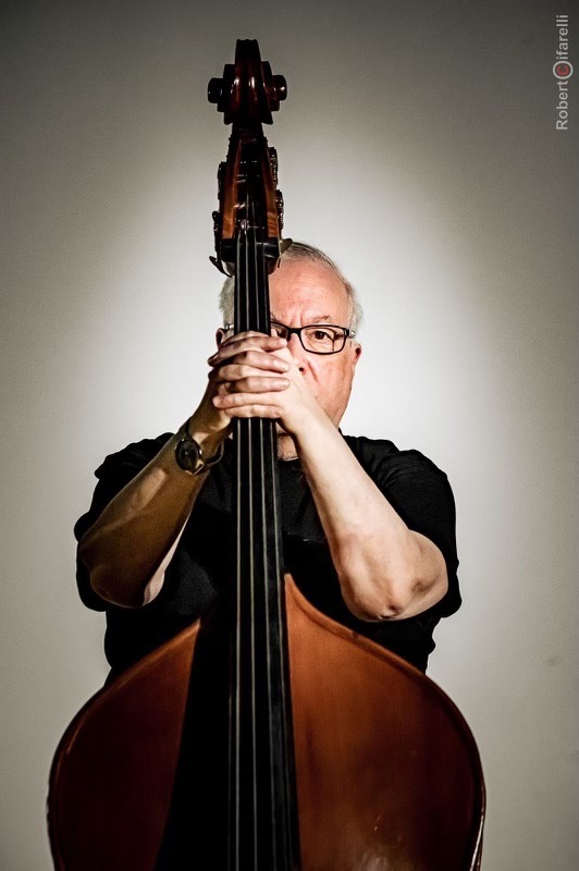 Portrait of a man holding an upright bass vertically in front of his face, standing against a neutral background, with only his eyes and arms visible around the instrument.