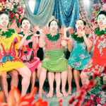 Five women in colorful dresses sit closely together in a vibrant, floral setting, posing expressively with bright makeup and animated facial expressions.