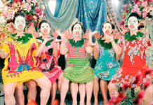 Five women in colorful dresses sit closely together in a vibrant, floral setting, posing expressively with bright makeup and animated facial expressions.