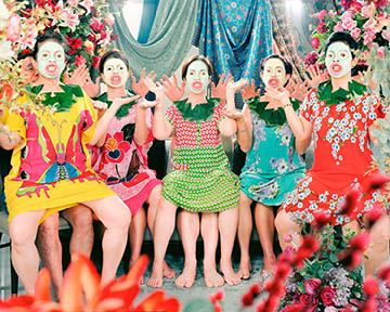Five women in colorful dresses sit closely together in a vibrant, floral setting, posing expressively with bright makeup and animated facial expressions.