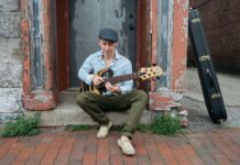 John Ferrara sits on brick steps outside a weathered doorway, playing an electric bass guitar. He wears a cap, patterned shirt, and casual shoes, focused on his instrument as he performs in a relaxed, urban setting.