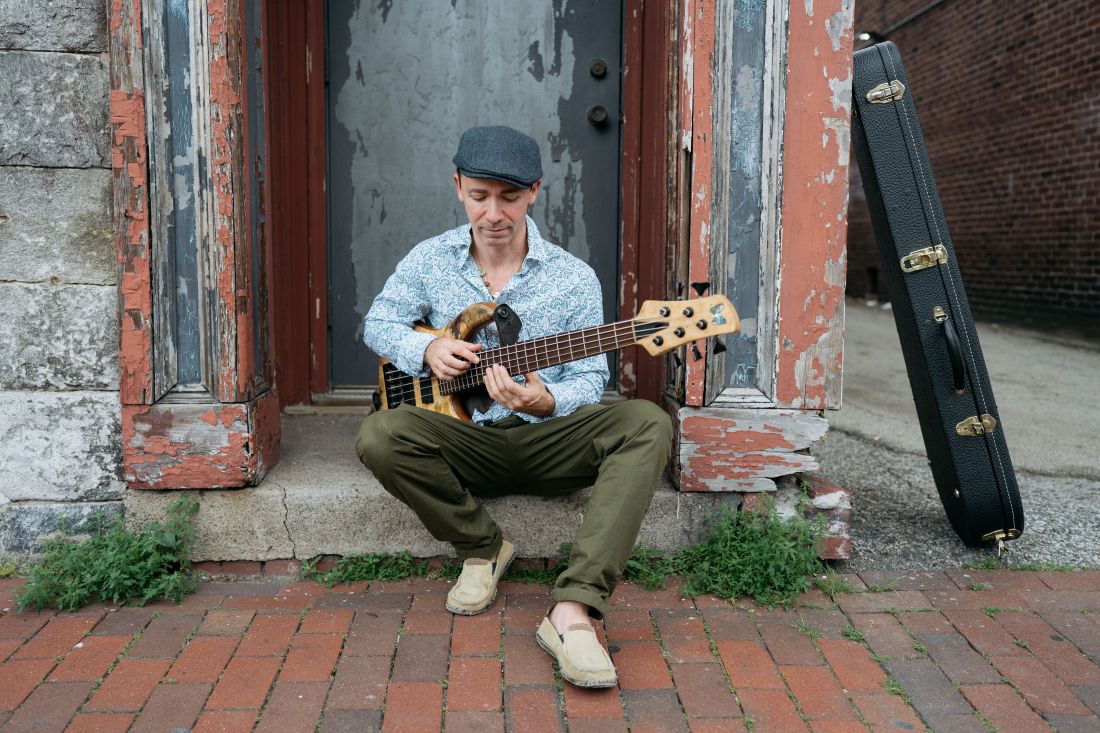 John Ferrara sits on brick steps outside a weathered doorway, playing an electric bass guitar. He wears a cap, patterned shirt, and casual shoes, focused on his instrument as he performs in a relaxed, urban setting.