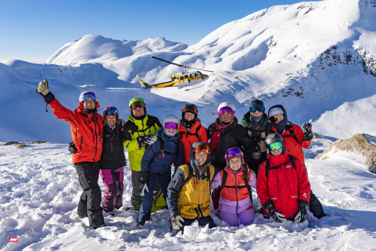 Skiiers pose at the top of a mountain while a helicopter flies in the background.