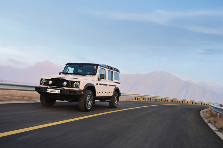 The INEOS Grenadier drives on a road in front of mountains.