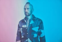 Portrait of a man standing against a soft blue and pink gradient background, wearing a patterned jacket over a light shirt. He faces slightly to the side with a calm, contemplative expression, illuminated by cool-toned lighting that enhances the ambient, atmospheric mood.