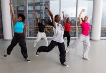 Group of young dancers practicing in a bright studio, mid-movement with arms raised and legs extended, wearing casual dance attire.