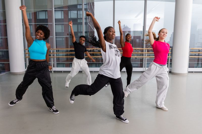 Group of young dancers practicing in a bright studio, mid-movement with arms raised and legs extended, wearing casual dance attire.