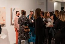 Guests mingle inside a contemporary gallery space during an exhibition opening, standing in small groups and chatting near displayed artworks on white walls. The atmosphere feels lively and social, with attendees dressed in smart casual attire and holding drinks while engaging in conversation.