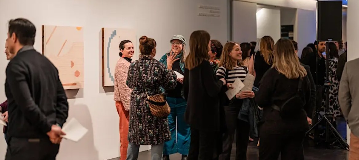 Guests mingle inside a contemporary gallery space during an exhibition opening, standing in small groups and chatting near displayed artworks on white walls. The atmosphere feels lively and social, with attendees dressed in smart casual attire and holding drinks while engaging in conversation.