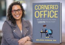Author Melissa Doman stands smiling beside a large display of her book Cornered Office: Why We Need to Talk About Leadership Mental Health. She wears glasses and a blazer, posing in front of the book cover, which features bold yellow lettering and an illustration of a professional seated at a desk, reinforcing the leadership and workplace theme.