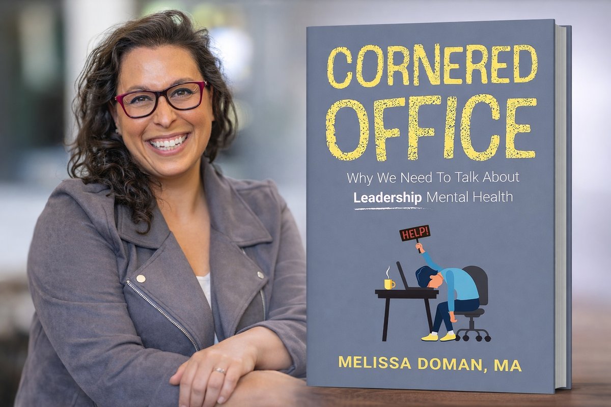 Author Melissa Doman stands smiling beside a large display of her book Cornered Office: Why We Need to Talk About Leadership Mental Health. She wears glasses and a blazer, posing in front of the book cover, which features bold yellow lettering and an illustration of a professional seated at a desk, reinforcing the leadership and workplace theme.