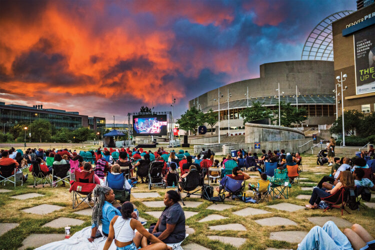 People watch a movie in a park with a sunset behind the movie screen.