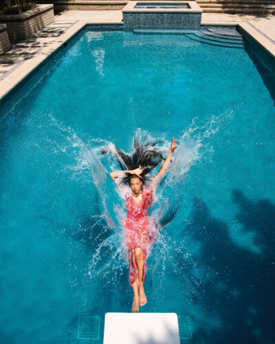 Woman falls into pool off of diving board while wearing floor length sundress.