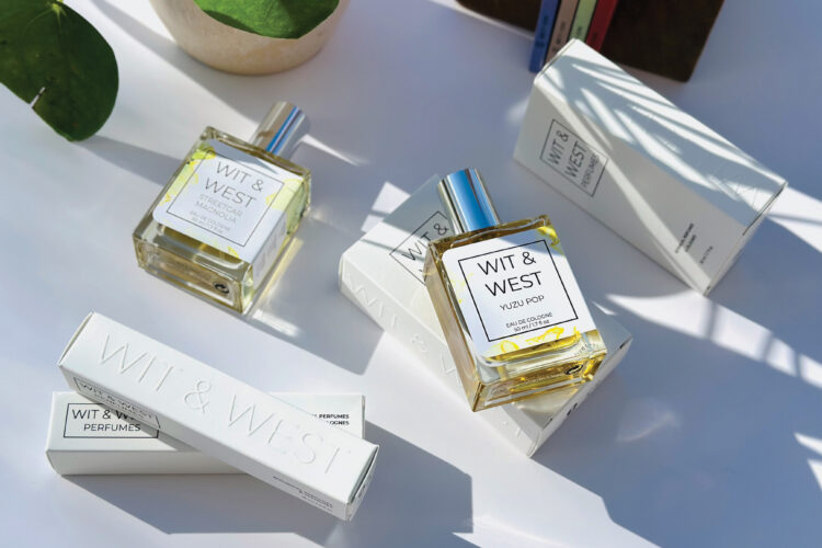 Wit & West perfume bottles and packaging arranged on a white surface in sunlight, featuring the Streetcar Magnolia and Yuzu Pop fragrances.