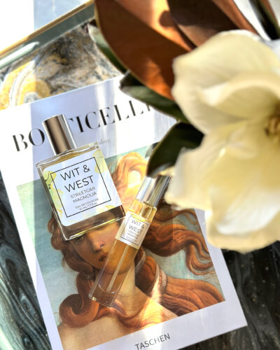 Wit & West Streetcar Magnolia perfume bottle and travel spray displayed on an art book beside a white magnolia flower.