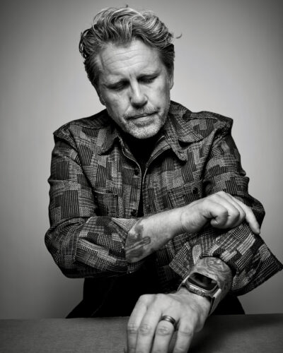 Black-and-white portrait of a man with wavy hair and a beard wearing a patterned jacket, seated with his arms crossed and looking downward.