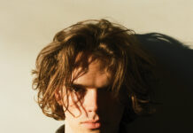 Highland Haircare Proves Clean Can Perform Portrait of a young man with tousled brown hair wearing a dark knit sweater, illuminated by dramatic sunlight and shadow.