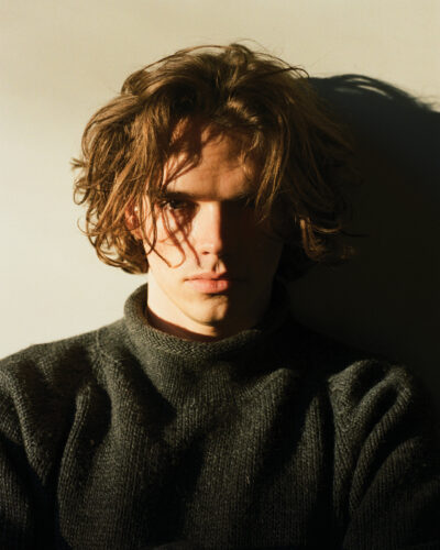 Portrait of a young man with tousled brown hair wearing a dark knit sweater, illuminated by dramatic sunlight and shadow.