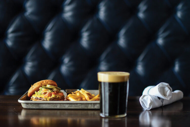 Burger, french fries, and a dark beer served on a table against a tufted leather booth.