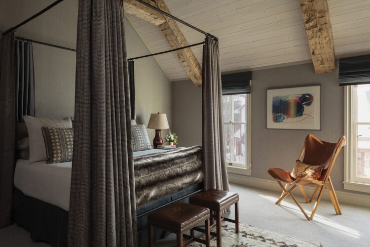 Cozy mountain guest room with a curtained canopy bed, exposed wood beams, leather chair, and framed artwork.