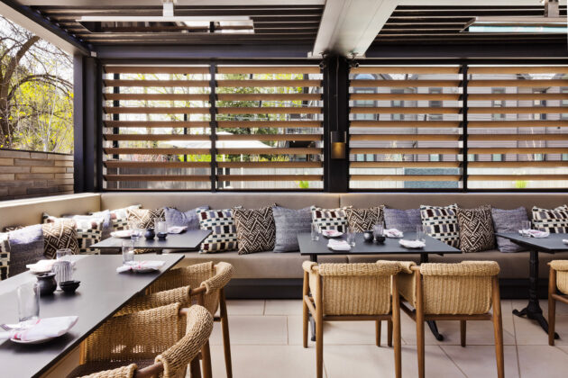 Bright restaurant patio with woven chairs, patterned cushions, dark tables, and wood privacy screens.