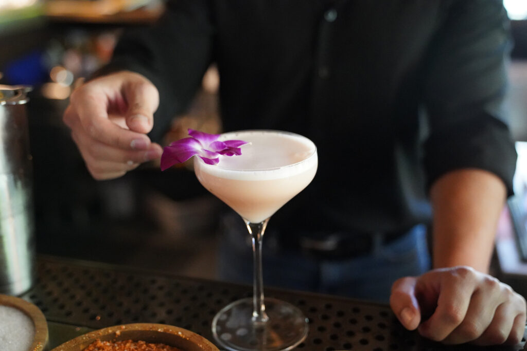 Bartender garnishing a pale pink cocktail with a purple orchid behind the bar.
