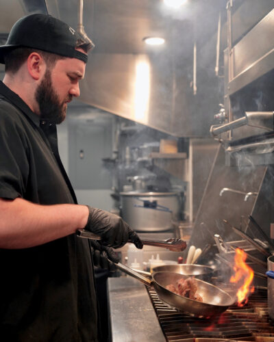 Chef cooking over an open flame in a stainless-steel restaurant kitchen.
