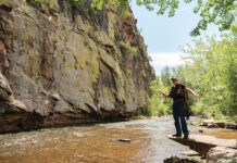 September Gets a Little More Rugged in Colorado Fly fisherman standing beside a rocky creek with fishing gear, surrounded by cliffs, trees, and summer greenery.