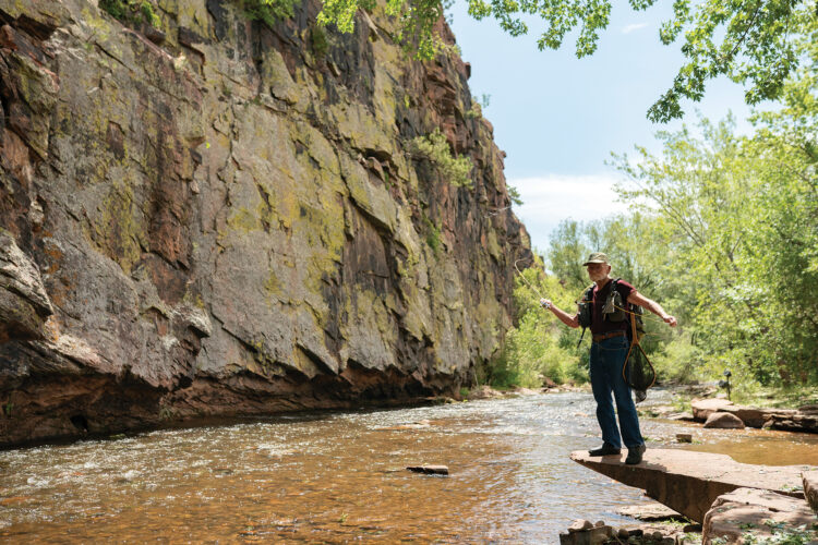 Fly fisherman standing beside a rocky creek with fishing gear, surrounded by cliffs, trees, and summer greenery.