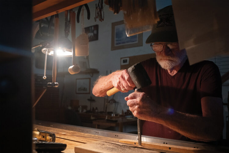 Craftsman working by lamplight in a workshop, using a mallet and hand tools to build a traditional bamboo fly rod.