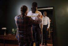 Tailor measuring a client’s shoulders for a custom shirt while the client looks into a mirror.