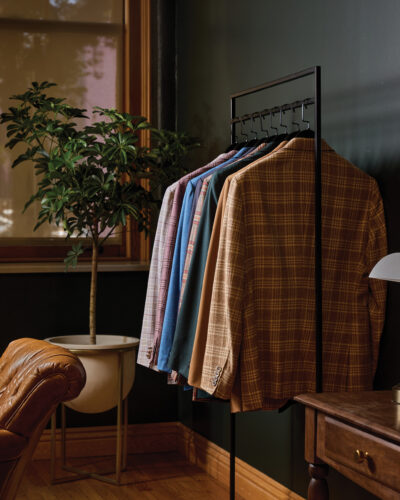 Collection of colorful plaid and solid suit jackets hanging on a clothing rack beside a potted plant and wooden furniture.