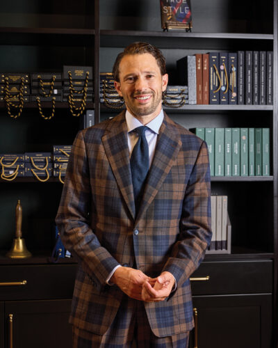 Menswear tailor wearing a brown-and-blue plaid suit, blue tie, and white dress shirt while standing in front of shelves filled with tailoring materials and books.