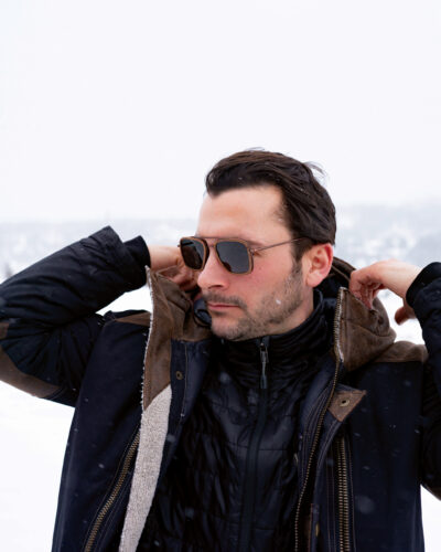 Man wearing sunglasses and layered winter outerwear while standing outdoors in a snowy mountain setting.