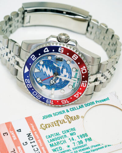 Stainless steel wristwatch with a colorful red-and-blue bezel and illustrated dial displayed beside a vintage Grateful Dead concert ticket.