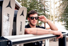 Man wearing sunglasses posing beside a pair of Atomic skis in a snowy Colorado mountain setting.