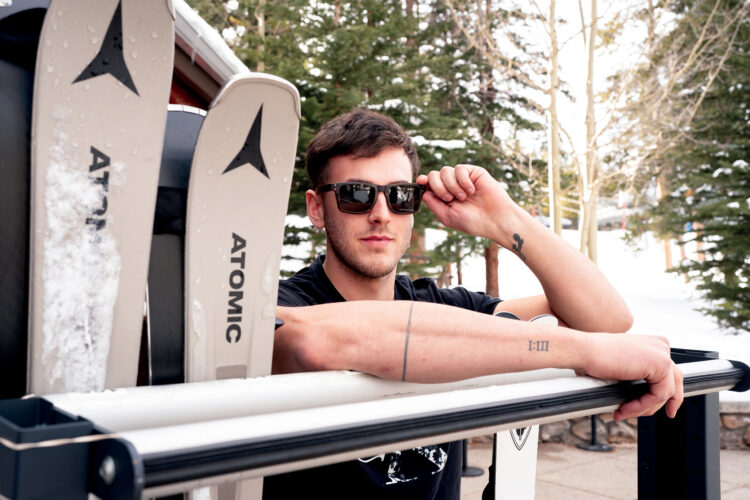 Man wearing sunglasses posing beside a pair of Atomic skis in a snowy Colorado mountain setting.