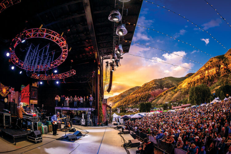 Live concert at an outdoor mountain venue, with musicians performing onstage before a large crowd as the sun sets over the surrounding peaks.