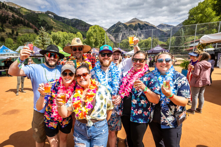 Group of festival goers wearing colorful shirts and leis and holding drinks at an outdoor beer festival surrounded by mountain scenery.