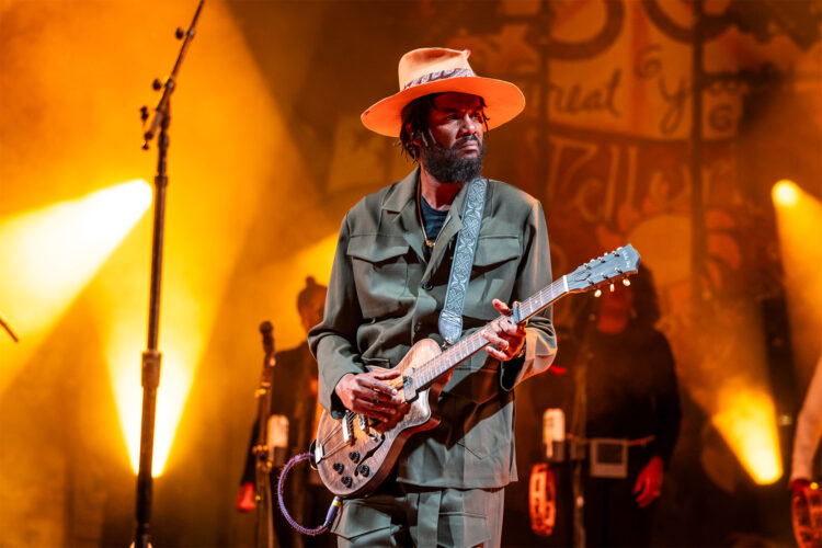 Guitarist performing onstage in a wide-brimmed hat and dark suit under dramatic golden concert lighting.