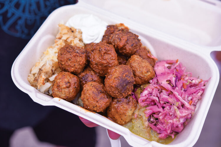 A takeout container filled with Ukrainian-style meatballs, potato pancakes, pink cabbage slaw, sauce, and sour cream.