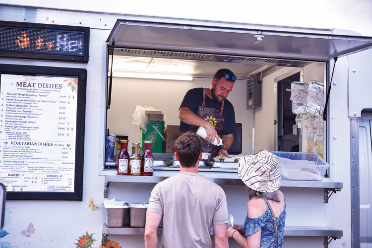 A vendor prepares an order inside the Sunflower Ukrainian Foods food truck as two customers wait at the service window.