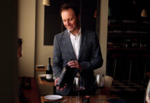 Scott Thomas poses in a plaid blazer presenting a bottle of red wine at a restaurant table with filled wine glasses.
