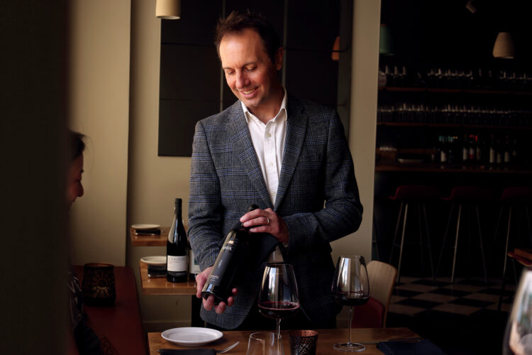 Scott Thomas poses in a plaid blazer presenting a bottle of red wine at a restaurant table with filled wine glasses.
