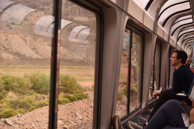Passenger looking through the panoramic windows of an Amtrak observation car as the train travels through a mountainous Western landscape.