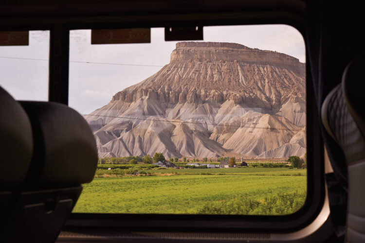 Rocky desert mesa and green farmland framed by the window of an Amtrak passenger train.